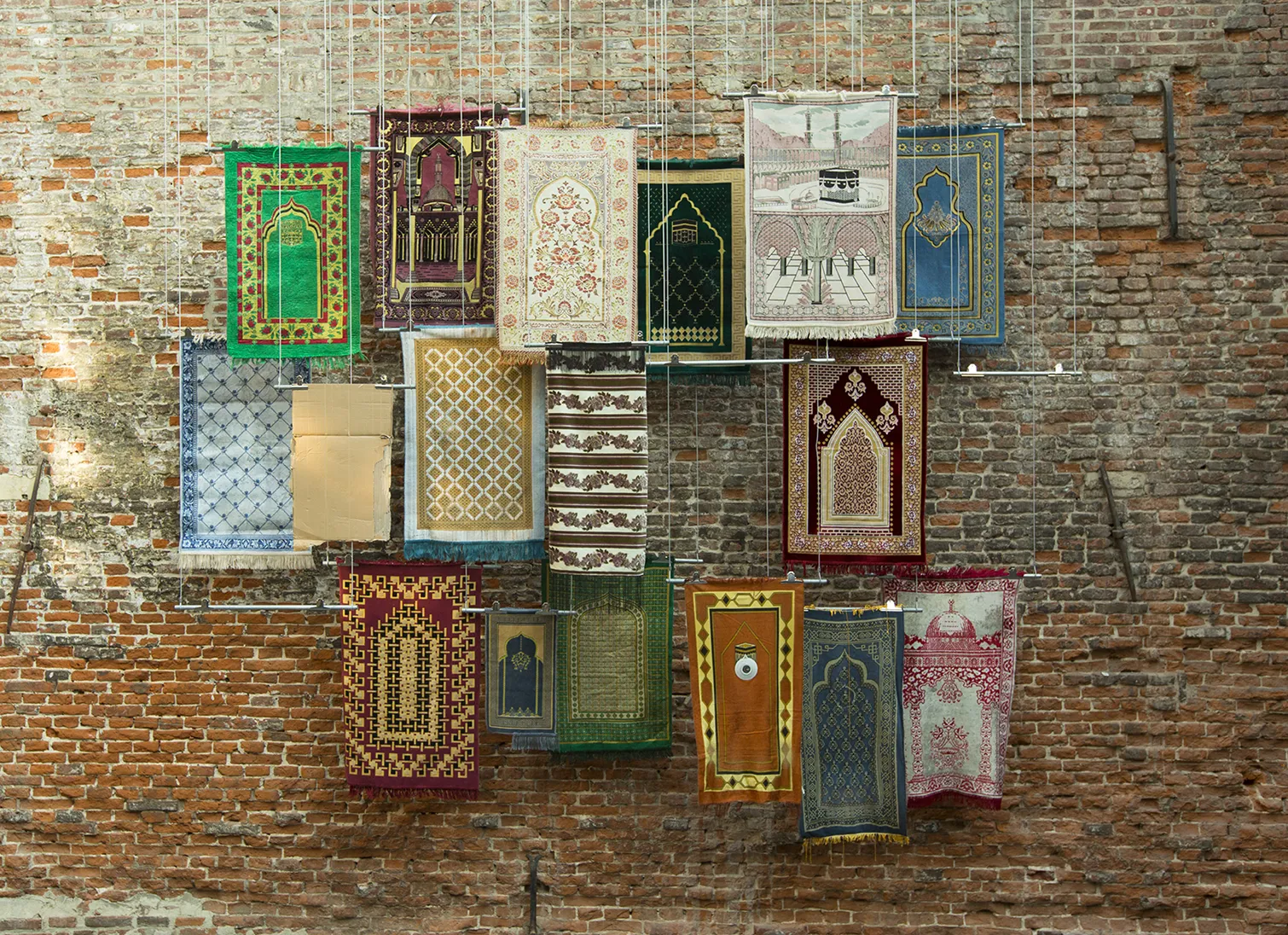 Photograph of an art installation featuring a selection of prayer rugs suspended in front of a brick wall.